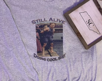 May include: A gray long-sleeve shirt with the text "Still Alive Doing Cool Shit" and a photo of a man in a boat.