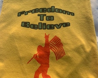May include: Yellow t-shirt with the text "Freedom To Believe" and a silhouette of a person holding an American flag.