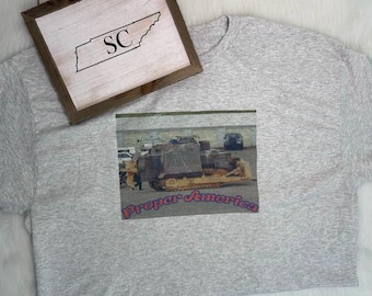 May include: A light gray t-shirt featuring a rectangular graphic print of a construction vehicle on a paved lot with the text Proper America in red and blue letters. Above the shirt sits a wooden framed sign with the outline of Tennessee and SC.