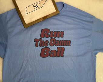 May include: A light blue t-shirt featuring the text Run The Damn Ball in dark red letters with a black outline. A wooden sign with a Tennessee state outline and the letters SC is positioned at the top left corner of the light blue t-shirt.