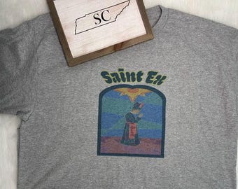 May include: A heather gray t-shirt featuring a graphic print with the text Saint Ex above an arched illustration of a figure in a dark robe standing in a landscape under a sun. A wooden sign with the outline of Tennessee and the letters SC is visible.