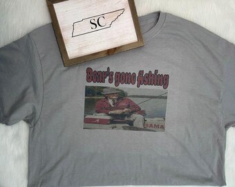 May include: A grey t-shirt featuring a graphic print of a person wearing a hat and red jacket while fishing on a boat. The text "Bear's gone fishing" is printed above the image. A wooden sign with the letters "SC" inside a state outline is visible.