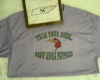 May include: A light purple t-shirt with the text "TRIM YOUR BUSH... GROW MORE PEPPERS!" in blue and yellow. The graphic features a red pepper with flames. A wooden sign with the outline of Tennessee and the letters "SC" rests on top.