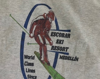 May include: A gray t-shirt with a blue and white graphic of a skier on a ski slope. The graphic includes the text "Escobar Ski Resort Medellin" and "World Class Lines Since 1978".