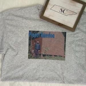 May include: A light gray t-shirt with a graphic that reads "Proper America" above an image of a person standing in front of a building. A framed wooden sign with the letters "SC" is in the upper right corner.
