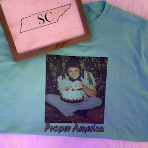 May include: A light blue t-shirt with a graphic of a person holding a cake, with the text "Proper America" in purple. A wooden framed sign with the outline of Tennessee and the letters "SC" is on top.