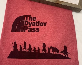 May include: Red t-shirt with a black graphic of a mountain range and the text "The Dyatlov Pass". Below the text is a silhouette of a group of people walking, including a person riding a horse.