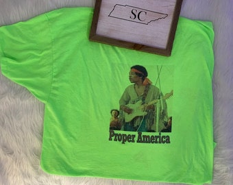 May include: A neon green t-shirt with a graphic of a man playing a guitar. The graphic is in a vintage style and includes the text "Proper America".