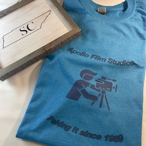 May include: A blue t-shirt with the text "Apollo Film Studios" and a graphic of a person holding a camera. The text "Faking it since 1969" is below the graphic.