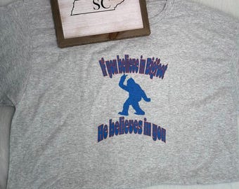 May include: A heather gray short-sleeved t-shirt featuring a blue silhouette of Bigfoot in the center. The text "If you believe in Bigfoot" is printed in blue above the figure, and "He believes in you" is printed in blue below the figure.