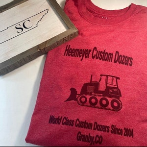 May include: Red t-shirt with a black graphic of a bulldozer and the text "Heemeyer Custom Dozers", "World Class Custom Dozers Since 2004", and "Granby, CO".