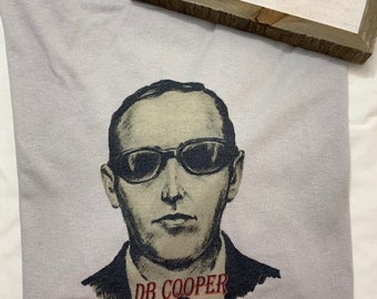 May include: A gray t-shirt with a black and white illustration of a man wearing sunglasses. The text "DB COOPER HIDE & SEEK CHAMPION" is printed in red on the shirt.