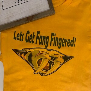 May include: Yellow t-shirt with a green and black graphic of a saber-toothed cat with the text "Let's Get Fang Fingered!"