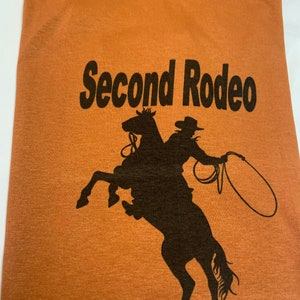 以下が含まれることがあります： 黒文字で「Second Rodeo」とプリントされた茶色のTシャツ。テキストの下には、馬に乗ったカウボーイの黒いシルエットがプリントされています。