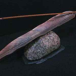 May include: A copper-toned feather-shaped incense holder with a brown incense stick. The holder is placed on a grey stone and a small black slate base, against a black backdrop. The incense stick is lit, with smoke rising.