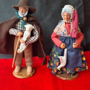 May include: Two ceramic figurines of a shepherd and a shepherdess. The shepherd is wearing a brown hat, a brown coat, and a plaid shirt. He is holding a staff and a white lamb. The shepherdess is wearing a white bonnet, a red dress, and a blue skirt. She is holding a white goose and a red umbrella.