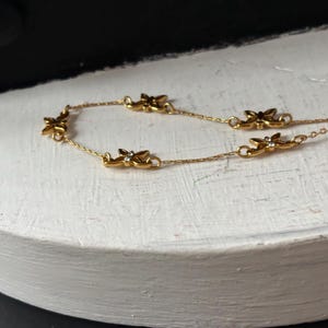 May include: A delicate gold-toned chain bracelet featuring small butterfly charms. Each butterfly has a small, clear gemstone in the center. The bracelet is displayed on a white, rounded surface against a dark background.