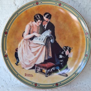 May include: A decorative plate featuring a couple reading a book, with a dog at their feet. The plate has a gold rim with green and red accents. The artwork is signed "Norman Rockwell". The background is a warm, golden hue.