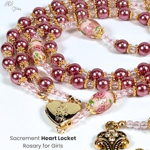 May include: A close-up of a rosary with pink beads and gold accents. The rosary features a heart-shaped locket and decorative glass beads with floral designs. The text "Sacrament Heart Locket Rosary for Girls" is visible.