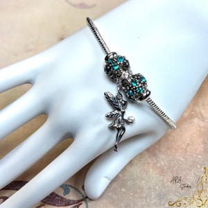 May include: A silver-toned snake chain bracelet featuring a dangling fairy charm holding a small white bead. The bracelet includes a decorative barrel-shaped bead encrusted with multiple small, bright teal-colored crystals set in silver metal.