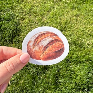 May include: A round sticker with a white background and a detailed illustration of a loaf of sourdough bread.