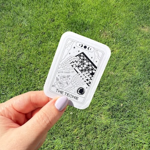 May include: A white sticker with a black and white illustration of skeleton hands typing on a keyboard. The sticker reads "NEVER LOGGED OFF" and "THE TECHIE". The sticker is held against a background of green grass.