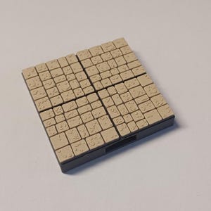 Op de afbeelding: Een set van 36 beige vierkante tegels met reliëfmotieven, gerangschikt in een 6x6 raster. De tegels zijn gemonteerd op een zwarte basis.