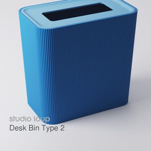 Pode incluir: Um caixote de secretária azul com uma abertura retangular e um exterior texturizado e canelado. O caixote tem uma borda azul clara e o texto "studio loup Desk Bin Type 2" é visível na lateral.