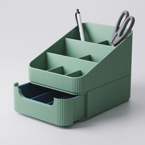 May include: A sage green desk organizer with multiple compartments and a pull-out drawer. A white pen and gray scissors are in the top section. The organizer has a ribbed texture and is designed for office or home use.