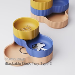 Pode incluir: Conjunto de bandejas de mesa empilháveis em amarelo, azul e pêssego. As bandejas têm uma textura canelada e são projetadas para se encaixarem. Uma bandeja contém clipes de papel, outra chaves. O texto "studio loud" e "Stackable Desk Tray Type 2" são visíveis.