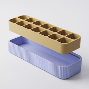 Puede incluir: Un juego de dos contenedores. La parte superior es una bandeja de color amarillo mostaza con 12 compartimentos cuadrados. La parte inferior es una bandeja de color lavanda con detalles acanalados verticales. Para almacenamiento.