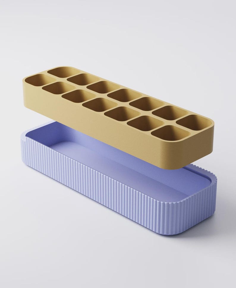 Puede incluir: Un juego de dos contenedores. La parte superior es una bandeja de color amarillo mostaza con 12 compartimentos cuadrados. La parte inferior es una bandeja de color lavanda con detalles acanalados verticales. Para almacenamiento.