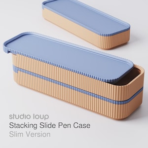 Könnte beinhalten: Zwei stapelbare Stifteetuis mit hellblauen Deckeln und beigefarbenen, geriffelten Seiten. Der Text "studio loup Stacking Slide Pen Case Slim Version" ist links unten aufgedruckt.