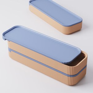 Puede incluir: Dos fiambreras rectangulares con tapas azul claro y laterales beige corrugados. Una caja est&aacute; abierta, revelando el interior. Una fina franja azul acent&uacute;a los lados. Las cajas est&aacute;n sobre un fondo blanco.