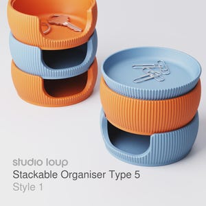 Puede incluir: Organizador apilable en naranja y azul. El organizador tiene una textura acanalada y una abertura curva. Una pila tiene una llave y un anillo, la otra clips. El texto "studio loup Stackable Organiser Type 5 Style 1" es visible.