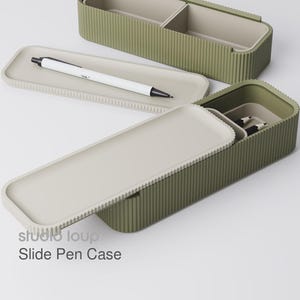 Peut inclure: Trousse &agrave; stylos coulissante vert olive avec int&eacute;rieur et couvercle gris clair. Un stylo blanc avec des d&eacute;tails noirs repose sur un plateau gris clair. Le texte "studio loup Slide Pen Case" est visible.