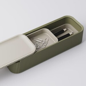 Peut inclure: Une trousse &agrave; crayons rectangulaire verte avec une texture stri&eacute;e, ouverte pour r&eacute;v&eacute;ler un plateau avec des trombones argent&eacute;s et deux crayons noirs. Le couvercle est blanc cass&eacute; et a une texture stri&eacute;e assortie.