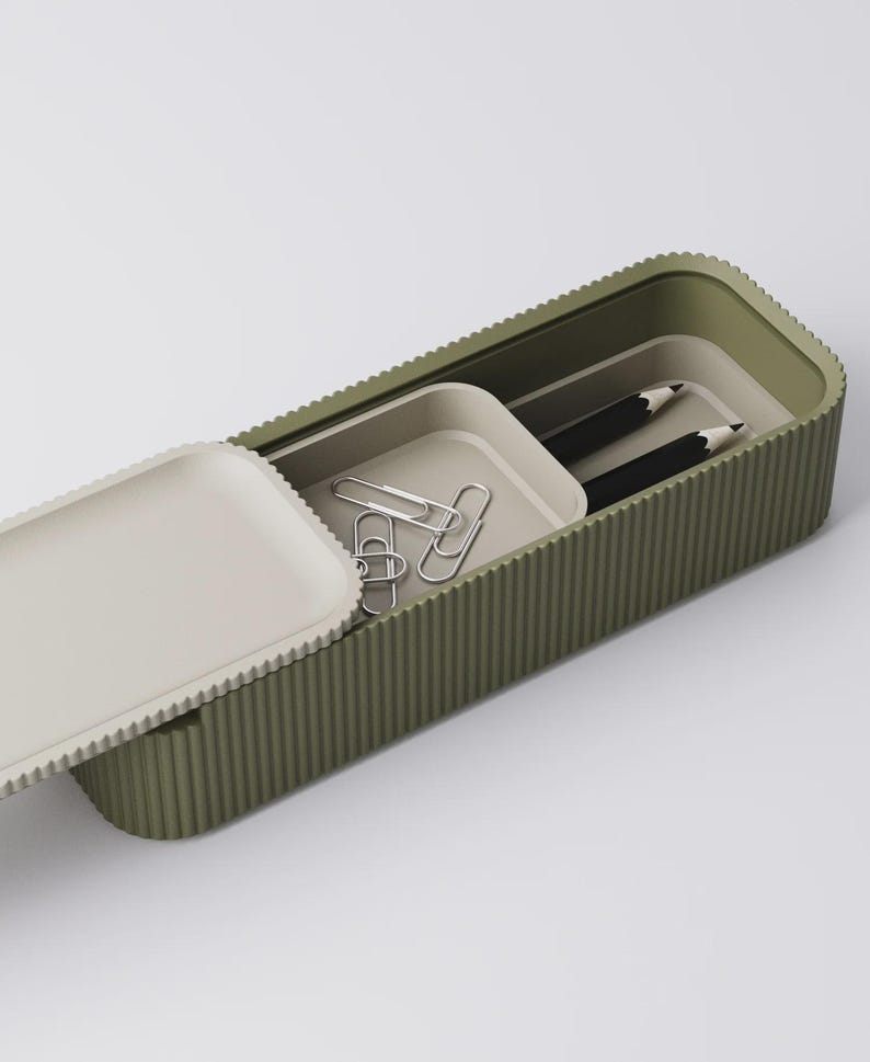 Peut inclure: Une trousse &agrave; crayons rectangulaire verte avec une texture stri&eacute;e, ouverte pour r&eacute;v&eacute;ler un plateau avec des trombones argent&eacute;s et deux crayons noirs. Le couvercle est blanc cass&eacute; et a une texture stri&eacute;e assortie.