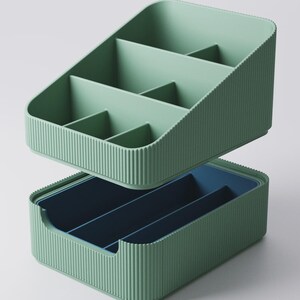 May include: Two-piece organizer set in a muted green color. The top section has multiple compartments, while the bottom section is a darker blue. Both pieces have a ribbed texture on the exterior, providing a modern aesthetic. Ideal for desk or vanity organization.