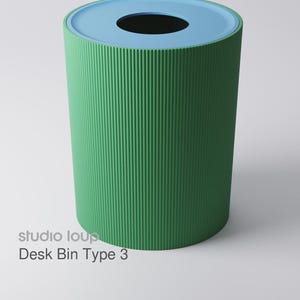 Könnte beinhalten: Ein grüner Schreibtischbehälter mit gerippter Textur und hellblauem Rand. Der Behälter hat eine zylindrische Form mit einer kreisförmigen Öffnung oben. Der Text "studio loup Desk Bin Type 3" ist auf der Seite aufgedruckt.