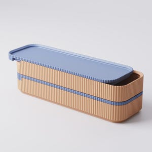 Puede incluir: Una fiambrera rectangular con un exterior corrugado marr&oacute;n claro y una tapa azul claro. La fiambrera tiene una banda azul claro alrededor del centro. La tapa est&aacute; parcialmente abierta, revelando el interior.