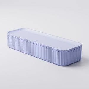Puede incluir: Una caja de almacenamiento rectangular de color lavanda con una tapa a juego. La caja tiene detalles verticales acanalados en los lados, creando una apariencia texturizada. La tapa es lisa y plana, con esquinas redondeadas.