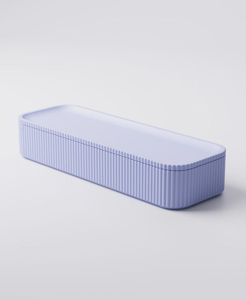 Puede incluir: Una caja de almacenamiento rectangular de color lavanda con una tapa a juego. La caja tiene detalles verticales acanalados en los lados, creando una apariencia texturizada. La tapa es lisa y plana, con esquinas redondeadas.