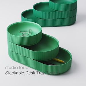 May include: Green stackable desk trays with a ribbed design. The trays are in a stepped arrangement, with a pencil and paperclips visible in the trays. The text "studio loup Stackable Desk Tray Type 0" is printed on the bottom tray.
