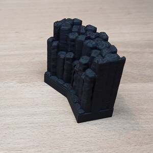 Puede incluir: Modelo negro impreso en 3D de una formación rocosa, posiblemente un acantilado o cañón. El modelo presenta estructuras verticales en forma de pilares y una superficie texturizada. La base es de forma rectangular oscura. El modelo se muestra sobre una superficie de madera clara.