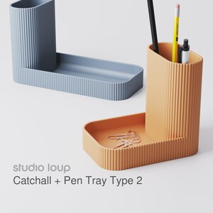 Peut inclure: Deux organiseurs de bureau en forme de L, bleus et oranges. L'organisateur bleu a un porte-stylos et un plateau. L'organisateur orange a un porte-stylos avec un crayon noir, un crayon jaune et un stylo, et un plateau avec des trombones. Le texte "studio loup Catchall + Pen Tray Type 2" est visible.