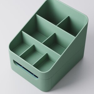 May include: A sage green organizer with multiple compartments. The rectangular container has a ribbed exterior and a handle on one side. The interior is divided into six sections, ideal for storing small items. The organizer is made of plastic.