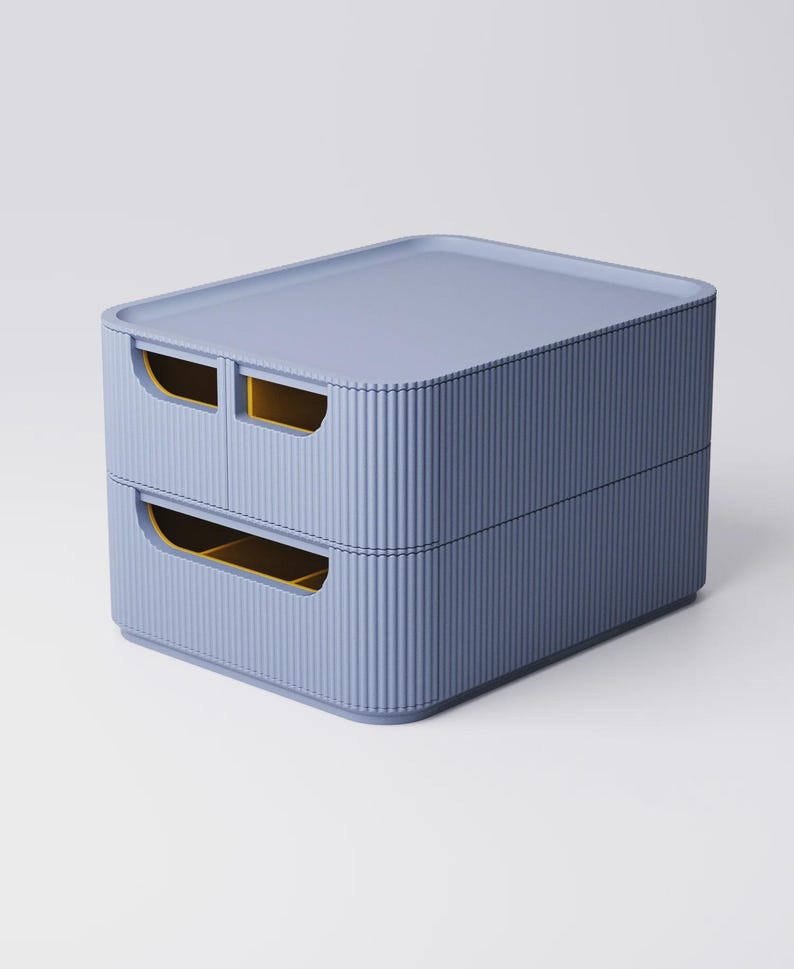 Op de afbeelding: Een opbergcontainer met twee lades in een gedempte blauwe kleur. De container heeft een geribbelde textuur aan de zijkanten en een platte bovenkant. De lade-openingen zijn rechthoekig met gele binnenkanten. Het ontwerp is modern en minimalistisch.