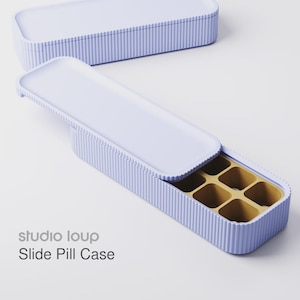 Op de afbeelding: Een lichtpaarse schuifpillendoos met een geribbelde buitenkant en een contrasterend gouden interieur. De doos is rechthoekig met een schuifdeksel. De tekst "studio loup Slide Pill Case" is zichtbaar.