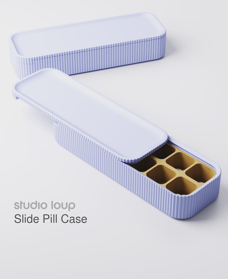Puede incluir: Un pastillero deslizante de color lila claro con un exterior acanalado y un interior dorado en contraste. La caja es rectangular con una tapa corredera. El texto "studio loup Slide Pill Case" es visible.